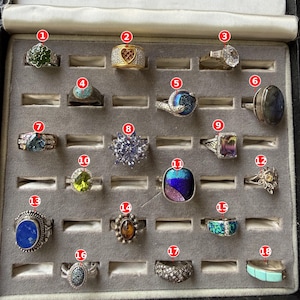 May include: A collection of eighteen assorted rings displayed in a gray velvet jewelry tray. The rings feature various gemstones including turquoise, amber, lapis lazuli, and clear crystals set in silver and gold-toned metal bands of different designs.