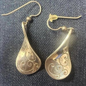 May include: A pair of silver-toned drop earrings featuring a teardrop-shaped pendant with an engraved swirling scroll pattern. Each earring is attached to a curved metal ear wire with a small bead accent and a clear plastic backing for support.