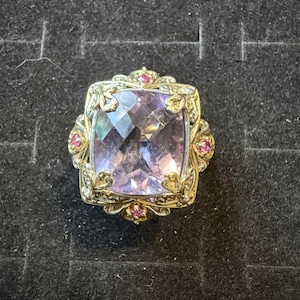 May include: A gold ring featuring a large, cushion-cut, light purple gemstone at the center. The ornate gold setting is decorated with small, round, vibrant pink accent stones placed at the top, bottom, and sides of the central gemstone.