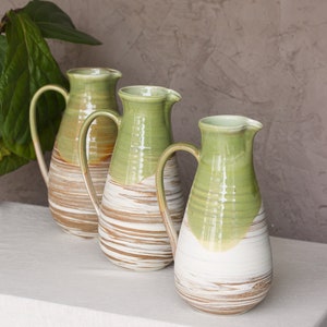 May include: Three ceramic pitchers with a green glaze and brown and white stripes. The pitchers are arranged in a row on a white tablecloth.