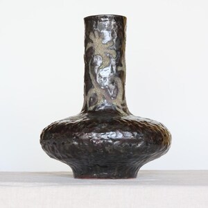 May include: A dark brown ceramic vase with a textured surface and a bulbous base. The vase has a tall, cylindrical neck with a lighter, patterned design. The overall shape is reminiscent of a chalice.