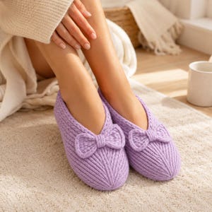 May include: A pair of light purple knit slippers featuring a ribbed texture and a decorative bow on the top of each slipper. The slippers have a rounded toe design and are crafted from a soft, textured yarn material with a slip-on style.