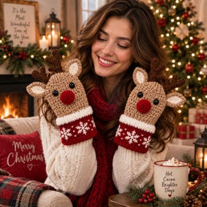 May include: A pair of tan crochet mittens featuring reindeer faces with dark brown antlers, black button eyes, and bright red noses. The cuffs are white with a red band decorated with white snowflake patterns, designed for winter wear.