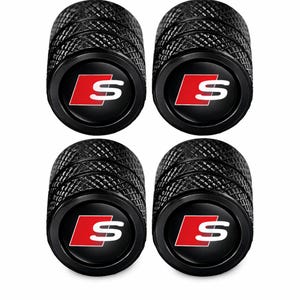 May include: A set of four black cylindrical tyre valve caps with a textured, knurled grip surface. Each cap features a circular emblem on the top displaying a red square with a white letter S, representing the Audi S-line logo design.