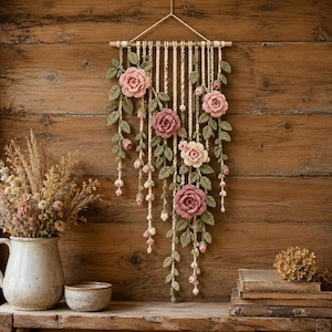 May include: A wall hanging featuring crocheted pink roses and green leaves suspended from a horizontal wooden dowel by cream-colored yarn strands. The floral arrangement includes multiple rose blossoms and small buds with detailed foliage.