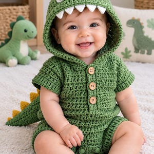 May include: A green crochet romper featuring a dinosaur hood with white teeth, yellow spikes, and black and white eyes. The garment has short sleeves, three brown wooden buttons on the front placket, and a matching green tail with yellow spikes.