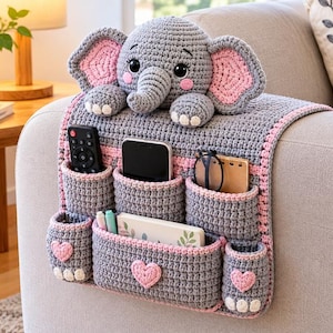 May include: A gray and pink crocheted sofa armrest organizer featuring an elephant head at the top. The organizer has multiple pockets holding a remote, smartphone, glasses, notebook, and pens. It is decorated with small pink heart appliques.