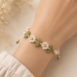 Può includere: Un braccialetto con motivi floreali intrecciati color crema e forme di foglie intrecciate verde oliva. Il design è accentuato da piccole perline metalliche color oro lucido intervallate tra gli elementi floreali e le foglie su un cordino crema.