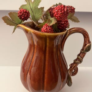 May include: A glossy brown ceramic pitcher with a vertical ribbed texture and a handle shaped like a twisted vine. The pitcher is filled with artificial green leaves and clusters of small, textured red berries that extend from the top opening.