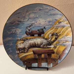 May include: A round decorative plate with a gold rim featuring a detailed illustration of a brown and white tabby cat sleeping on a yellow striped armchair. The scene includes a milk carton and a small box on a wooden side table against a cloudy sky.
