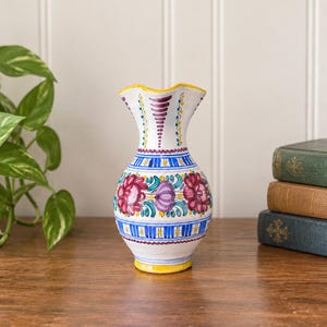 May include: A white ceramic vase with a flared, wavy rim and a rounded body. The surface is hand-painted with vibrant floral motifs in shades of magenta, purple, and teal, accented by horizontal blue and yellow stripes and yellow trim.