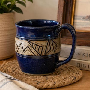 May include: A ceramic mug featuring a glossy deep blue glaze on the top and bottom sections, with a central band of unglazed, speckled beige clay decorated with incised geometric patterns, including triangles and lines, and a curved handle.