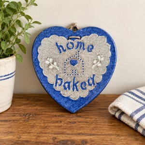 May include: A heart-shaped ceramic wall plaque with a glossy blue border and a textured beige center. The text "home baked" is embossed in blue, accompanied by a small blue heart icon and two white floral sprigs. A jute string loop is attached at the top.