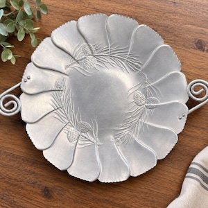 May include: A round silver-toned metal serving tray with a scalloped edge and two spiral handles. The surface features an embossed design of pine cones and pine needles arranged in a circular pattern around the smooth center of the tray.