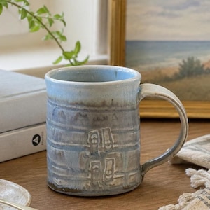 May include: A cylindrical ceramic mug with a light blue and grey glaze. The exterior features horizontal embossed lines and a repeating stamped pattern of small, rectangular architectural shapes. It has a sturdy, curved handle on the right side.