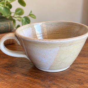 May include: A wide, shallow ceramic mug with a glossy, light beige glaze and a subtle white wash effect. The mug features a rounded handle on the left side, a flared rim, and a visible footed base, showcasing a smooth, hand-thrown pottery texture.