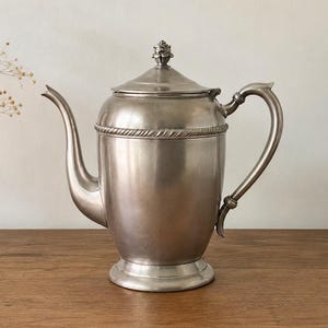 May include: A silver-toned metal coffee pot with a smooth, polished finish. The vessel features a curved spout, a sturdy handle, and a removable lid topped with a small, decorative finial. A rope-like embossed band encircles the upper body.