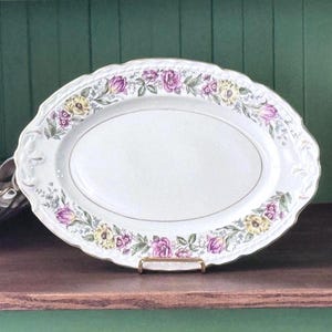 May include: An oval white ceramic serving platter featuring a decorative border of pink and yellow flowers with green leaves. The rim of the platter has a subtle embossed texture and a scalloped edge, framing a plain white center area.