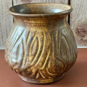 May include: A small, round ceramic vase with a wide, flared rim and a tapered base. The exterior features a textured, organic pattern of raised, swirling ridges finished in a mottled, earthy brown glaze with darker brown and golden highlights.