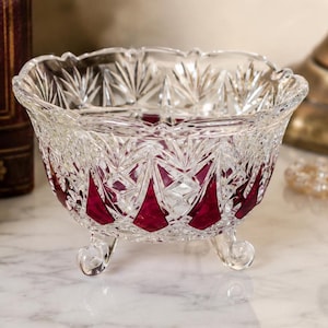 May include: A clear and cranberry red cut glass bowl featuring a complex geometric starburst pattern and a scalloped rim. The bowl is supported by three small, curved glass feet, showcasing a deep red color at the base of each faceted section.