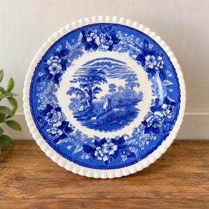 May include: A round ceramic plate with a scalloped edge, featuring a white center and a deep blue transferware pattern. The design depicts a pastoral landscape with trees and figures, surrounded by a wide border of detailed floral motifs.