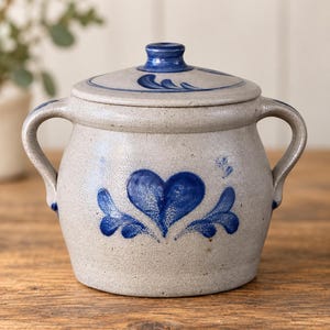 May include: A light gray stoneware crock with two side handles and a matching lid. The vessel features a hand-painted cobalt blue heart design on the front and blue decorative accents on the lid. The surface has a speckled, textured finish.