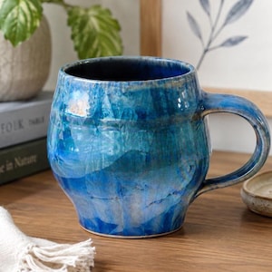 May include: A ceramic mug with a rounded, bulbous body and a curved handle. The surface features a glossy, multi-tonal glaze in shades of deep cobalt, sapphire, and lighter sky blue, creating a swirling, marbled effect across the entire exterior.