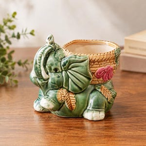 May include: A ceramic planter shaped like a green elephant with a tan woven-texture basket on its back. The elephant features a raised trunk, large ears, and a small pink flower detail on the side of the basket. The interior is cream-colored.