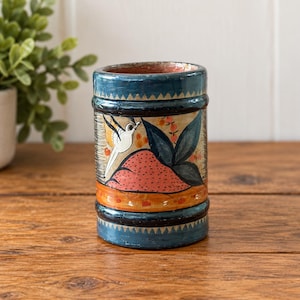 May include: A cylindrical wooden container featuring a hand-painted design with a blue border, a white bird in flight, a pink textured mound, and dark green leaves. The base and top rims are decorated with a dark blue and white triangular pattern.