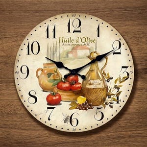 Puede incluir: Un reloj de pared redondo con esfera color crema, números negros y dos manecillas negras. El diseño muestra una escena pintada con una botella de aceite de oliva de cristal, una jarra de cerámica, tomates rojos y aceitunas con el texto Huile d'Olive.