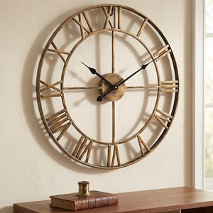 May include: A large, circular wall clock featuring an open-frame design with a metallic gold finish. The clock face displays black Roman numerals and two slender black hands. A central circular hub connects the frame, which is mounted on a wall.