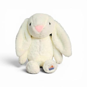 May include: A plush stuffed animal in the shape of a rabbit with cream-coloured fur, long floppy ears, and a small pink nose. The toy features a small rectangular tag on its left foot with a graphic of a bear and a chick and the text Stuffed.