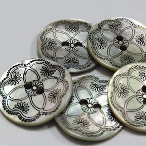 Może przedstawiać: Kolekcja pięciu okrągłych, iryzujących guzików z kwiatowym wzorem. Każdy guzik ma centralny kwiat i mniejsze kwiaty, wszystkie obrysowane na czarno. Guziki mają dwa otwory do szycia. Średnica około 2,5 cm.