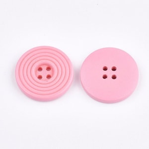 May include: Two pink wooden buttons with four holes each. One button has a concentric circle design.