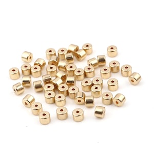Peut inclure: Un grand groupe de perles en métal de couleur dorée. Les perles sont rondes avec un trou au centre. Elles sont éparpillées sur une surface blanche.