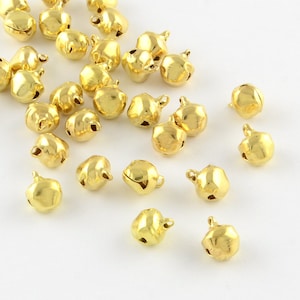 May include: A scattering of gold-colored metal bells with a loop at the top for hanging. The bells are scattered on a white surface.