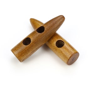 May include: Two wooden toggle buttons with a light brown finish are shown, each with three holes. The buttons are cylindrical with rounded ends and are positioned diagonally across each other against a white background. These buttons are ideal for coats and jackets.