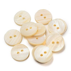 May include: A collection of round, off-white buttons with a pearlescent finish. Each button features two centered holes for attachment. The surface of these circular fasteners displays a subtle, natural grain pattern characteristic of shell material.