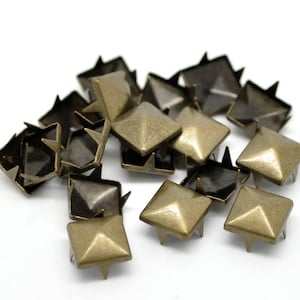 May include: A group of 15 antique gold-toned metal pyramid-shaped studs with four points on the bottom. The studs are scattered on a white surface.