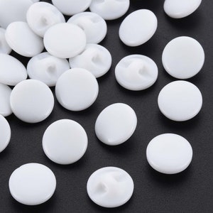 May include: A collection of white plastic buttons with a snap closure. The buttons are scattered on a black surface.