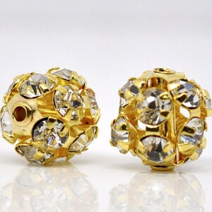 May include: Two gold-toned round beads with clear rhinestones. The beads have a hole in the center for stringing.