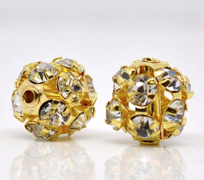 May include: Two gold-toned round beads with clear rhinestones. The beads have a hole in the center for stringing.