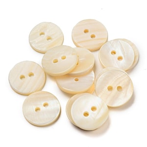 May include: A collection of round, off-white buttons with a smooth, iridescent finish. Each button features two centered holes for attachment. The surface of these circular fasteners displays a subtle, natural grain pattern with a glossy sheen.