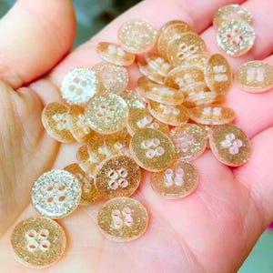 May include: A collection of small, round buttons with a glittery, gold appearance. Each button has four holes for sewing. The buttons are a light, translucent gold color, with the glitter adding sparkle. These buttons are ideal for sewing and craft projects.