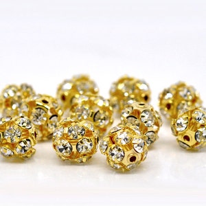 May include: A group of gold-toned round beads with clear rhinestones. The beads are scattered on a white surface.