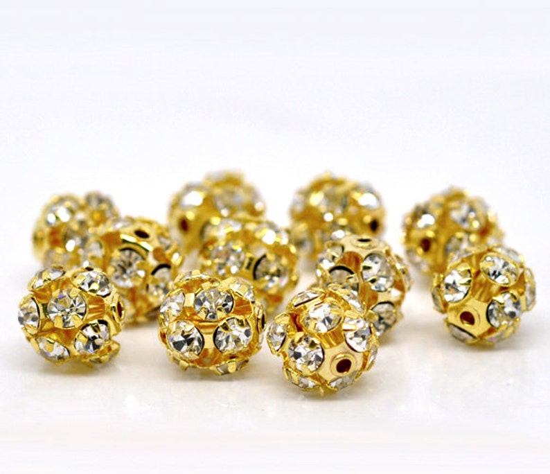 May include: A group of gold-toned round beads with clear rhinestones. The beads are scattered on a white surface.