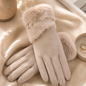 Könnte beinhalten: Ein Paar beigefarbene Handschuhe mit einem weichen Kunstfellbesatz und einem dekorativen Riemen mit einer goldfarbenen Metallschnalle. Die Handschuhe haben eine glatte, matte Oberfläche und sind mit einzelnen Fingerabschnitten gestaltet.