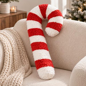 May include: A plush candy cane pillow featuring a textured, fuzzy surface with alternating horizontal stripes in bright red and white. The pillow is shaped like a curved hook, mimicking the classic appearance of a traditional holiday candy cane.