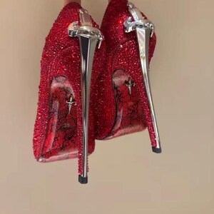 Puede incluir: Un par de zapatos de tacón alto rojos cubiertos de pequeños cristales rojos. Los tacones tienen forma de dagas metálicas de tono plateado. Las suelas presentan un patrón de encaje negro sobre un fondo rojo con un pequeño emblema de daga plateada y el número 36.