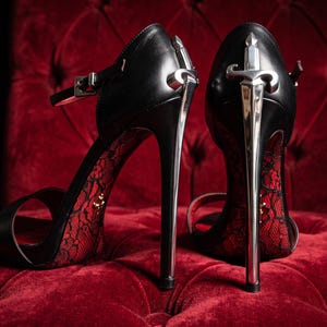 May include: A pair of black high-heeled sandals featuring a silver-toned stiletto heel shaped like a sword. The interior of the shoes is lined with a red lace pattern, and each shoe includes a thin ankle strap with a silver buckle closure.